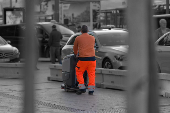 A street cleaner in an orange uniform is pushing a cleaning machine along the road, with taxis in the background. The image is monochrome except for the uniform. It appears to be shot through a fence or grid with blurry bars in the foreground.