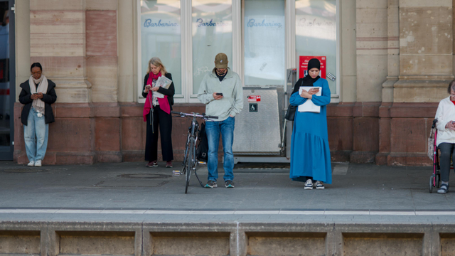 Five people, standing on a train platform, each of them for themselves, watching at their smartphones