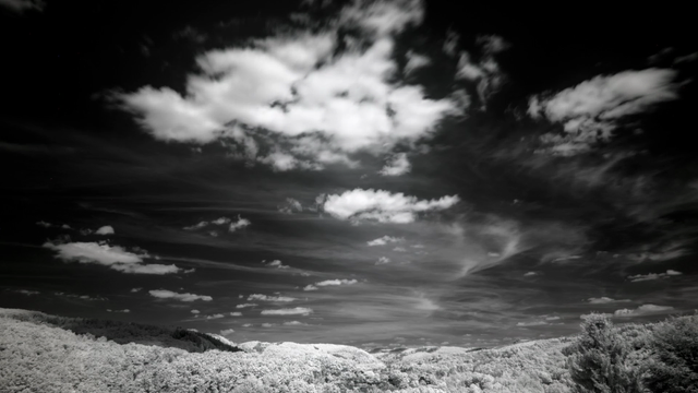 Monochrome infrared video of the sky above a hill line. The sky itself is black, with bright white clouds forming in the center of the image and moving closer to the viewer.