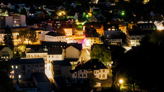 Night shot of the center of a small town from above, showing many two- and three-story houses with white and beige walls, and the streets between them, illuminated by lanterns and moving cars.