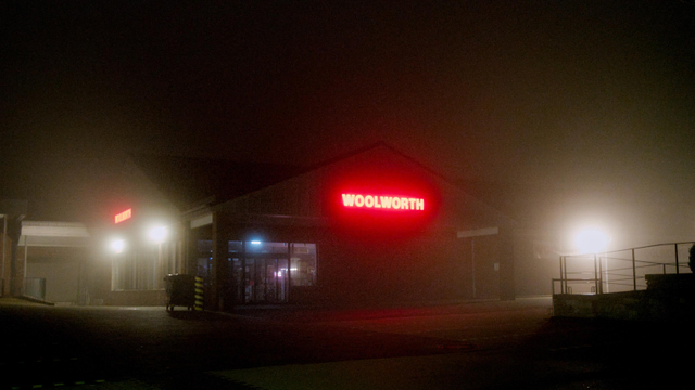 Night fog photo of a store front. To the left and to the right are bright white spotlights, illuminating the delivery ramp and the parking lot. In the middle, the red company logo "Woolworth" is showing a spooky red aura.