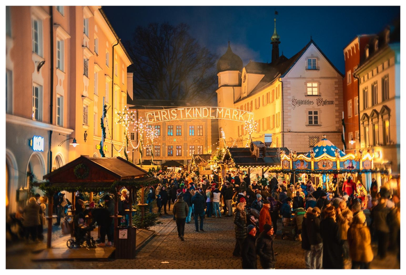 Bild entlang einer Straße, darüber hängt der leuchtende Schriftzug "Christkindlmarkt". 