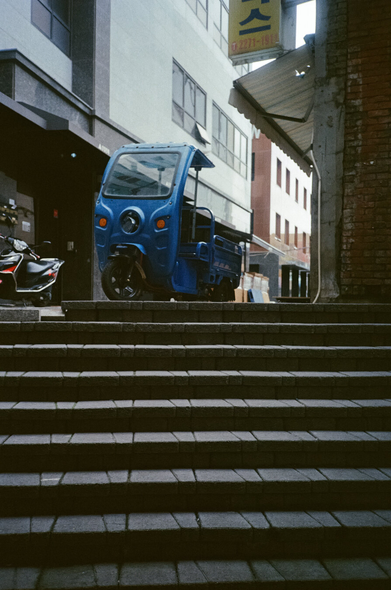 계단 위에 파란색 삼륜차 한 대가 정차해있다. 앞 부분에는 차창이 있다.