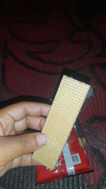 Close-up of a person's hand holding a rectangular wafer biscuit with a textured, grid-like pattern. The biscuit is light brown, and the hand appears to be of a medium skin tone. Visible in the background are a red package and a patterned, maroon surface.