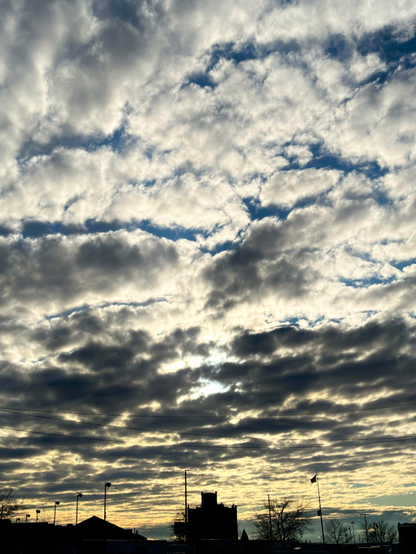 Photo of a sky filled with low clouds on a winter afternoon, over some silhouettes of buildings and streetlights at bottom frame. Some sunlight breaks through the cloud cover over a castle-like silhouette in the center, and the sky has a touch of blue and gold closer to the the horizon.