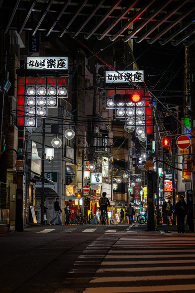 A man on a motorbike is entering a dark street at night passing through a gate lit with red and white art deco streetlamps, in Namba, Osaka, Japan. The scene is bathed in a cyber punk atmosphere.