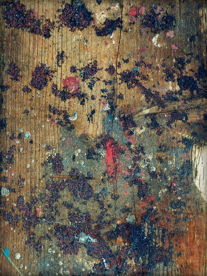 A colour photograph showing a random spattering of paint on a piece of shipwright's wood. The spatter resembles an explosion, seemingly emanating from a single point in purples, blues and red.