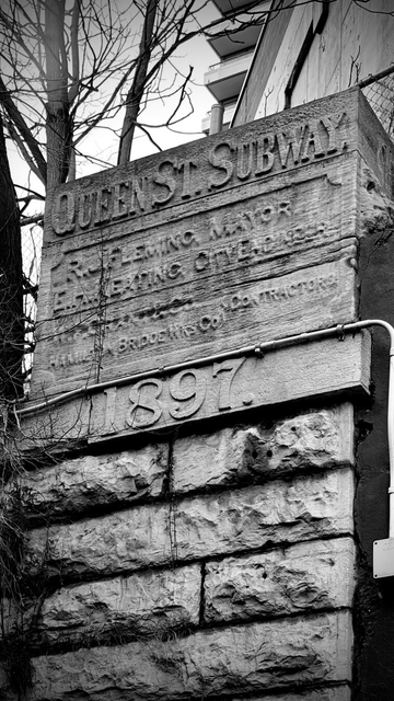 Raised sign carved on the face of a stone “Queen Street Subway” and “1897.” are the most visible words; the remainder are eroded names. The sign is one face of a stone block, atop of a large column of rough hewn stones set like bricks that act as a bridge support and retaining wall. Behind and above the sign, some trees, a bit of sky, and the edge of a multistorey apartment building are visible. Photo is black-and-white although the day was fairly grey so it was already pretty much black and white before photo alteration.