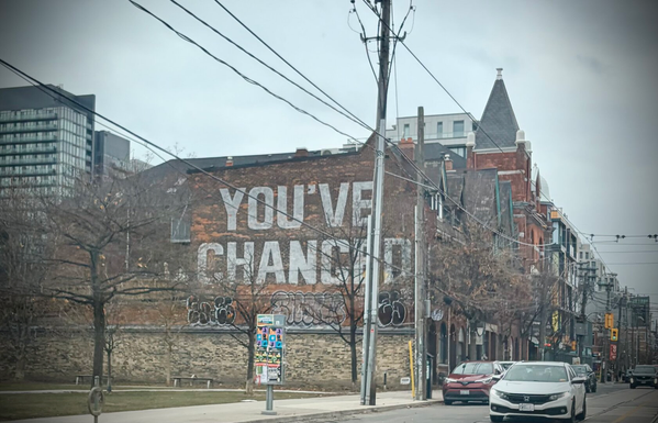 Mural on the side of a red/brown brick building says “you’ve changed” in all capital letters in white paint. There’s a low retaining wall in front of the building that faces into a park area. A few graffiti tags are visible below the mural. On the left you can see some glass and concrete skyscrapers, and on the right is the streetscape of older 2-3 story brick buildings. The foreground is both trees and hydro poles with wires. It’s a grey wintery day, although no snow is visible.