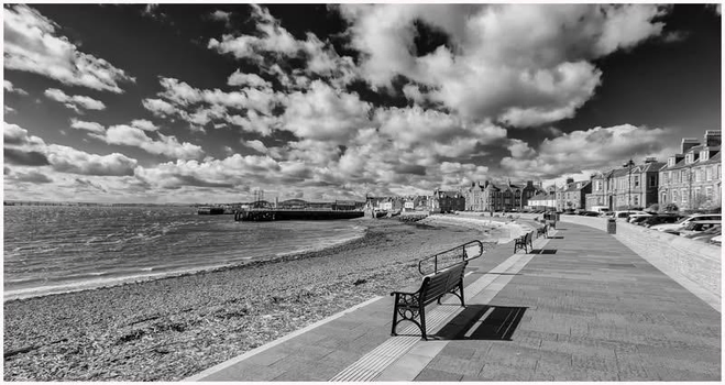 A black and white image of a seaside promenade featuring benches along the edge of a pebbled beach. In the background, there are buildings lining the waterfront and a pier extending into the water, with a dramatic sky filled with clouds.