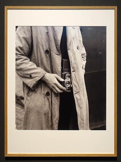 Zwart-wit foto die laat zien hoe de Nederlandse fotograaf Cas Oorthuys in Amsterdam tijdens de hongerwinter in 1945 stiekem foto's maakte van de verschrikkelijke taferelen in de hoofdstad. De camera hing onder een lange regenjas die makkelijk kon worden geopend om snel een foto te maken.