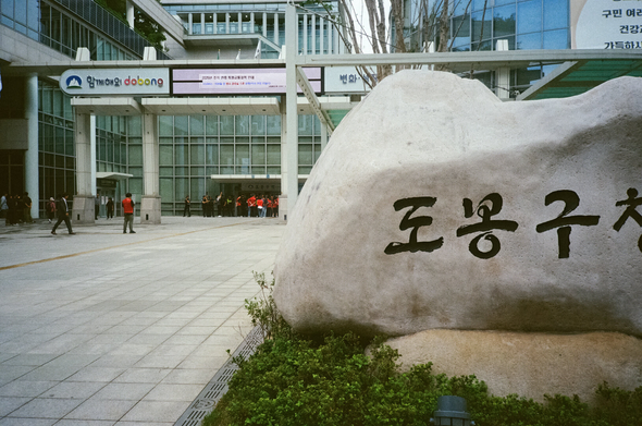 '도봉구청'이라 새겨진 거대한 바위 뒤로 화려한 도봉구청 입구가 보인다. 입구 앞에서 면담 약속을 어긴 도봉구청장을 직접 만나겠다는 노조 조합원들과, 이를 가로막고 있는 경찰·공무원들이 실랑이를 벌이고 있다.