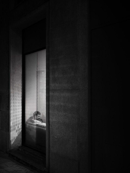 A young man sits on a sofa inside a window at night. Soft light comes out of the window while the rest of the scene is dark.