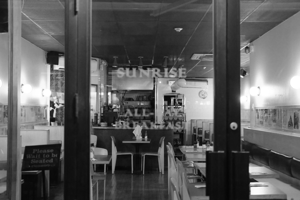 Night. Looking in through the windows of a closed restaurant. The interior is dim, the place settings laid out for the next day. Wall sconces light the waiting tables. A sign reads “Please wait to be seated.” The words on the door are slightly out of focus, only partially visible: “Sunrise. All…. Breakfast.”