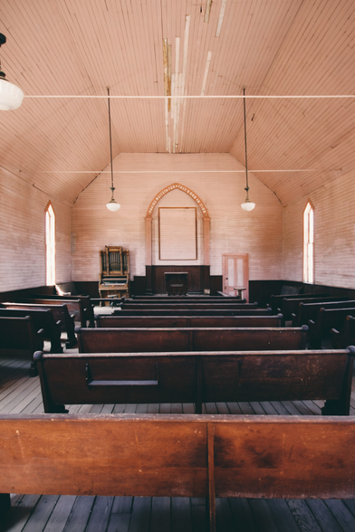 Photograph of the spartan inside of a small and abandoned wooden church in a desolate American mining town. The white paint on the walls and ceiling has browned, slowly approaching the hues of the dark wooden pews and altar. The text ‘Praise waiteth for thee O God in Zion’ has been painted in white on a wooden arch above the altar, and beside it stands a very small organ.