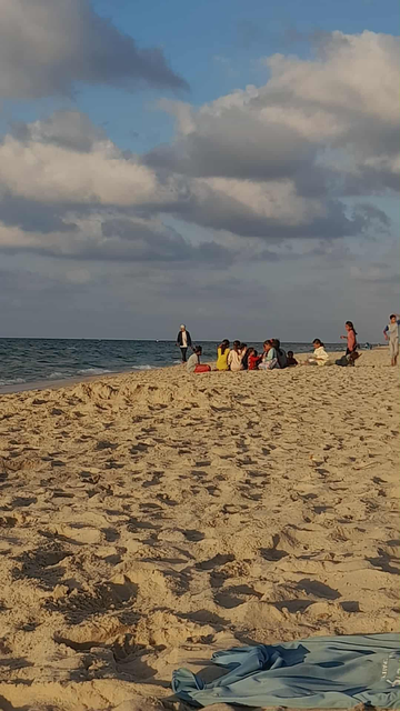 Picture of the beach in Gaza Stripe, in the south. We can see blue sky with clouds, a bit of the sea and lot of sand. Some people are enjoying the beach in the background.