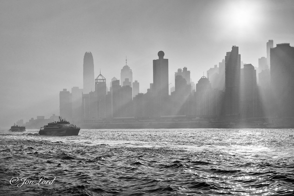 This is a black and white cityscape photo of the rising sun's sunbeams shining through the high-rise waterfront overlooking Victoria Harbour, Hong Kong (2018).

The lower quarter of the photo is filled with dark and choppy water with small wavelets of about one metre breaking the water's surface. To the left of centre are two ferries, one SeaCat fast ferry viewed nearly in profile and underway from left to right. The other, a standard (slow) ferry, a little more distant and underway from right to left and nearing the left margin. Above the waterline and forming an urban horizon, is a city waterfront filled nearly entirely with high-rise apartment and office blocks, all viewed in silhouette with little fine detail to be seen. The sky above is an early morning tropical haze, with a visibility of just a few km. In the upper right corner is a newly risen sun, just above the skyscrapers. It's brilliant light causing multiple shadows where the high-rise towers block the sunlight. Between these shadows are numerous sunbeams shining through the castellated cityscape behind and onto the water below.

The location: Victoria Harbour and the cityscape is the waterfront of Central District on Hong Kong island.