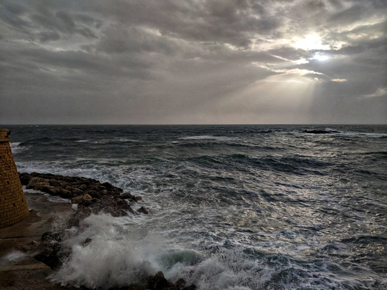 A dramatic seascape captured at dusk, featuring a choppy, dark grey sea with white waves crashing against a rocky shoreline. The sky is filled with thick, moody clouds, allowing a burst of sunlight to break through near the horizon, casting a golden glow over the water. On the left side of the image, a section of an old stone or brick breakwater is visible, leading the eye towards the vast expanse of the ocean. The scene evokes a sense of raw natural beauty and tranquillity amidst the powerful movement of the waves.