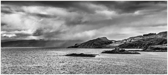 A black and white landscape image featuring a calm body of water with rocky outcrops and hills in the background. The sky is overcast with dramatic cloud formations.
