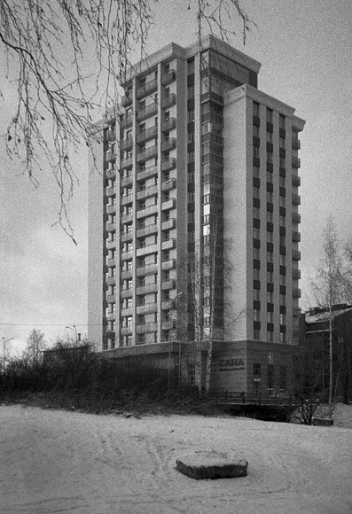 The black-and-white photo shows a high-rise apartment building. There is snow in front of the building, bushes and a dark manhole sticking out from under the snow are visible. In the foreground, branches of a tree hang from behind the frame.