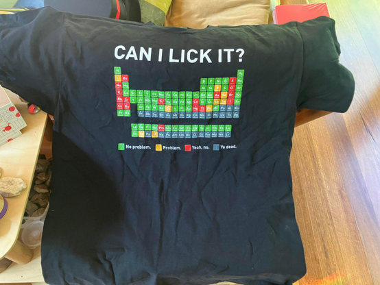 A black t-shirt with the periodic table printed on it. The elements have been colour coded to answer the question “Can I lick it?” The legend is: Green = No problem, Yellow = Problem, Red = Yeah, No, Dark grey = Ya dead.