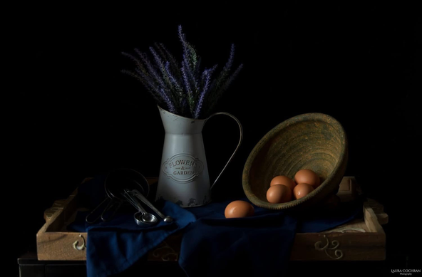 A subtly lit traditional still-life with: a bowl with eggs, a pitcher with flowers, and measuring cups and spoons on a tray with cloth. 