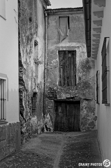 A narrow alleyway featuring aged, weathered walls and wooden doors, flanked by buildings with rustic textures. The scene is depicted in black and white, emphasizing the historical charm and character of the architecture.
