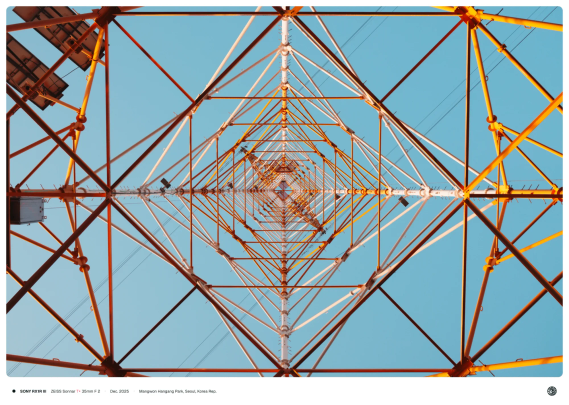 주황색 전신주의 아래 정 중앙에서 위를 바라보며 찍은 모습, 철골이 마름모꼴 모양의 패턴으로 쌓여 하늘로 올라가있다.