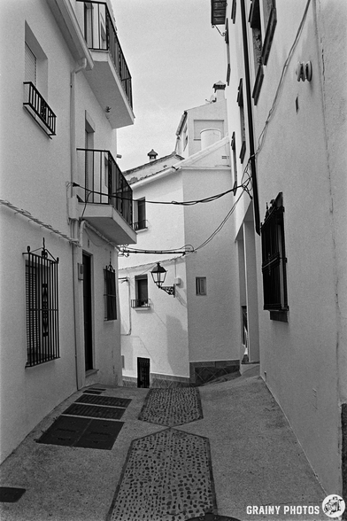 A narrow, winding alleyway lined with white buildings, featuring balconies and a street lamp, creating a charming and serene atmosphere. The cobblestone path adds texture to the scene, typical of quaint Spanish village architecture.
