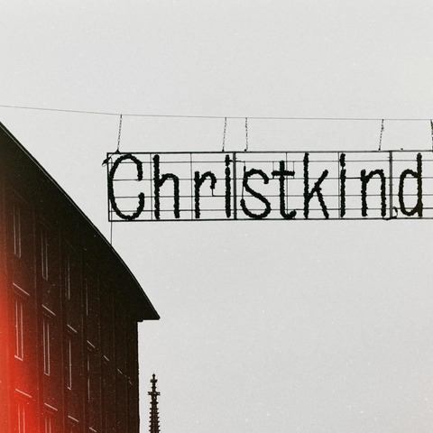 a black and white image of a christmas decoration hanging across the street. the sign uses the german name "christkindlesmarkt" to welcome visitors to the christmas market. in this cropped version only the part "christkind" of the sign is visible meaning christ child