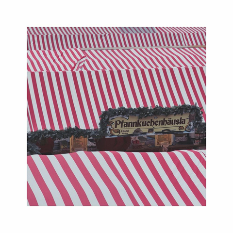 overlooking the textile roofscape of a christmas market. the textiles are painted in red and white stripes. ine of the aisles between the various huts becomes visible. the image is focusing on a decorative sign with the german word "pfannkuchenhäusla" meaning pancake house