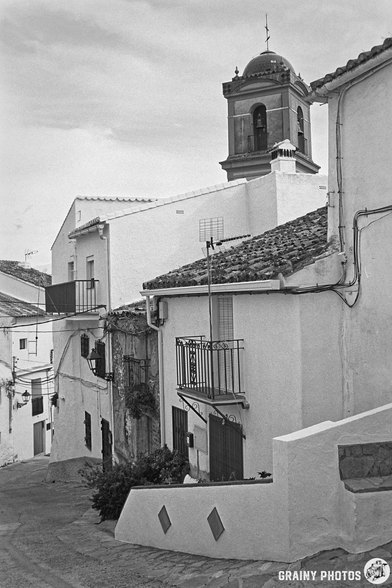 A narrow, winding street in Algatocín framed by whitewashed houses with balconies, leading up to a bell tower. The scene is captured in black and white, highlighting the charming architecture and cobblestone details.