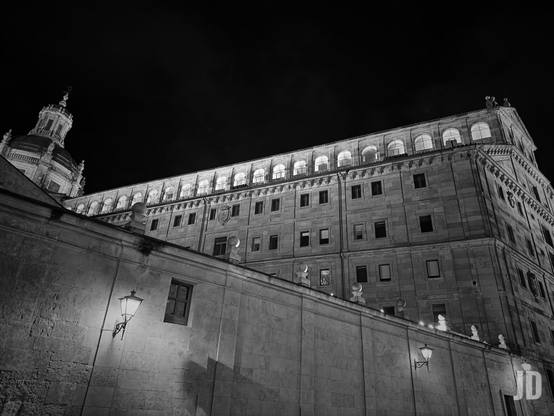 Imagen en blanco y negro de un imponente edificio de piedra iluminado por la noche, tomada desde un ángulo bajo mirando hacia arriba. En primer plano, un robusto muro de piedra con una farola antigua que proyecta luz sobre su superficie texturizada y pequeños elementos decorativos en la parte superior. Detrás del muro, se eleva el edificio principal de múltiples pisos, con numerosas ventanas rectangulares y una hilera de ventanas arqueadas brillantemente iluminadas en el piso superior. A la izquierda, se distingue una cúpula ornamentada con un remate. El cielo nocturno es completamente oscuro, creando un fuerte contraste con la arquitectura iluminada.