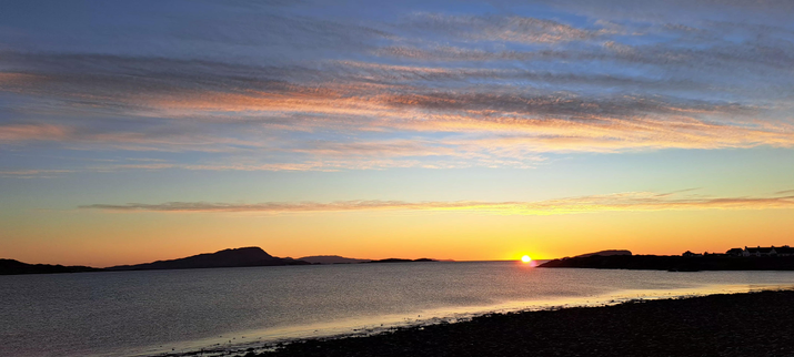 The sun sets in the Hebrides; a calm sea,  a sky that graduates from warm orange to icy blue holding a few clouds; dark silhouettes of islands that are between a few hundred metres away and 50 kilometres.