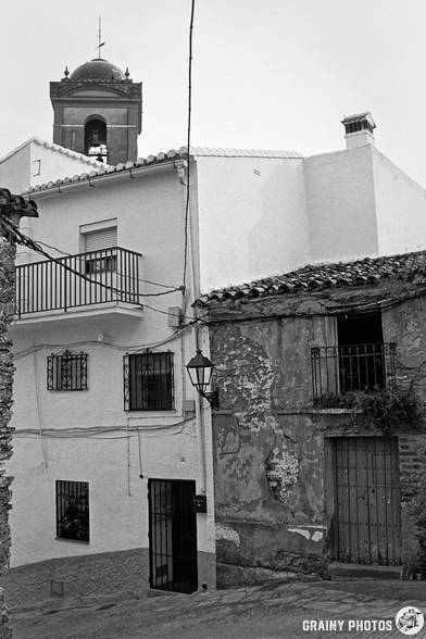 A black-and-white image of a quaint street scene featuring historic buildings. The architecture includes a mix of whitewashed exteriors and textured stone walls, with a prominent bell tower in the background and decorative window grilles.