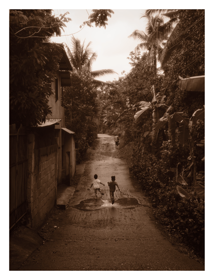 Two young children running down a wet, narrow road in a lush, rural setting. Flanked by simple houses on one side and dense tropical vegetation—including palm and banana trees—on the other, the children appear to be splashing through puddles as they run toward a third figure walking in the distance. - Google Gemini 3 Pro Preview