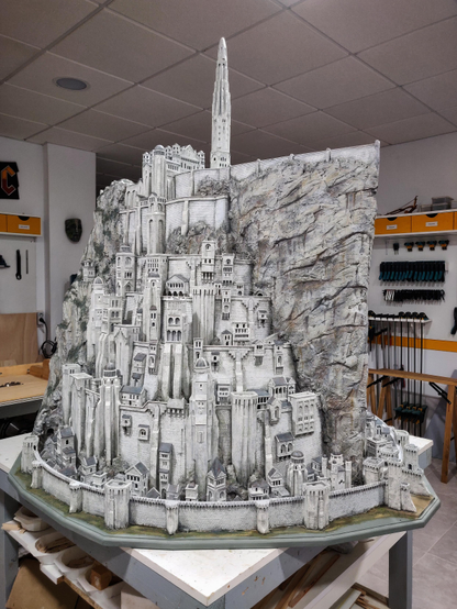 A model of a tall fortress city. Buildings are arranged in different circular levels, each smaller than the one below, like a wedding cake. All levels are crowded with lots of different buildings and towers of various sizes, giving the city a busy look. On the top level there's a palace and a tall spire. There's a huge flat rocky spur slicing the city in half. The buildings and rocks are mostly white. The model rests on a table, with some tools visible in the background.