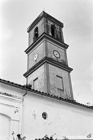 A black and white photograph of a historic church tower with clock and a bell, featuring a weathered exterior, situated against a clear sky above a white church wall.