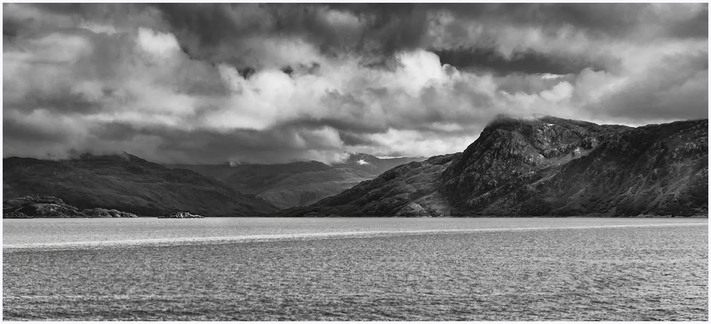 A black and white landscape featuring a calm body of water in the foreground, bordered by rugged hills and mountains. Dramatic clouds fill the sky above, creating a moody atmosphere.
