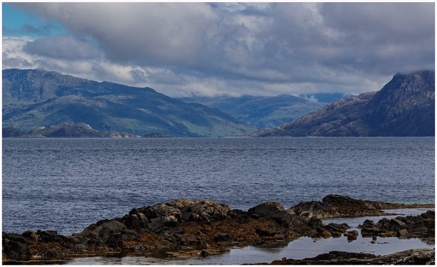 A scenic view of a calm sea with rocky shorelines in the foreground, surrounded by lush green hills and mountains under a partly cloudy sky.
