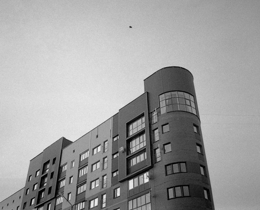 The black-and-white photo shows a multi-storey building of modern architecture. A bird flies over the house.
