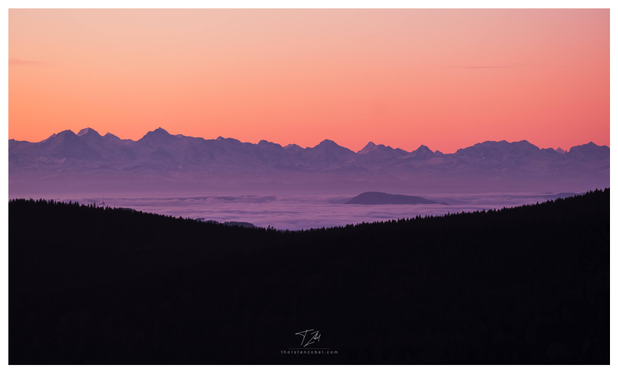 The lower part of the image is dominated by a dark, wooded chain of hills. Behind it, a hill rises out of a blanket of clouds. In the background, a mountain range with snow-capped peaks can be seen, above which the sky is colored orange-red by the rising sun.