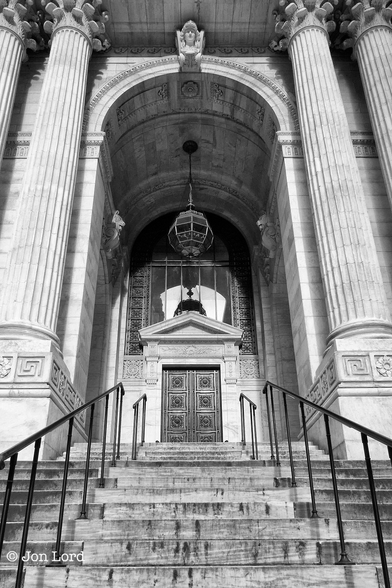 This is a black and white street photo in portrait format of the stairs leading up to the ornate entrance to the 42nd Street Library in New York City (2012).

From the base of the photo and rising upwards and away from the camera is a set of stone steps, twelve in view, with a simple black metal handrail on either side. These steps lead to large and ornate entrance. On the sides are four large and tall stone, perhaps marble, columns of about ten metres high. Between the columns is an arched and covered entrance with a single large metal and glass light suspended from top of the arch. At the entrance is a relatively small, about three metres high, black metal, double door. Above the door are large rectangular window panes rising up to the top of the stone arch.

The location: the 42nd Street Library, sometimes called the New York Public Library Main Branch or the New York Public Library or the Stephen A. Schwarzman Building in mid-town Manhattan NYC - A great building and it was a Sunday.