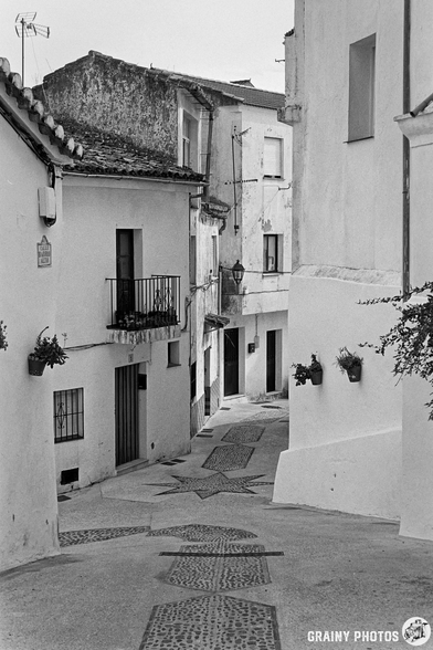 A narrow, cobblestone street meanders through Algatocín, lined with houses featuring balconies and flower pots. The scene captures the quiet atmosphere of a historic area, evoking a sense of nostalgia and tranquility.