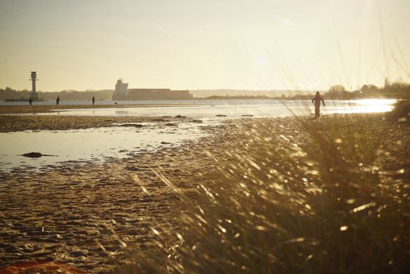 Nachmittägliche Sonne über dem Falckensteiner Strand. Links der Leuchtturm, mittig ein zugefrorener Priel, rechts ist ein Kind durch Dünengras sichtbar 