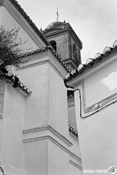 A black and white photo capturing the corner of a building with textured walls and a distinct bell tower in the background, framed by roof edges and overgrown foliage.