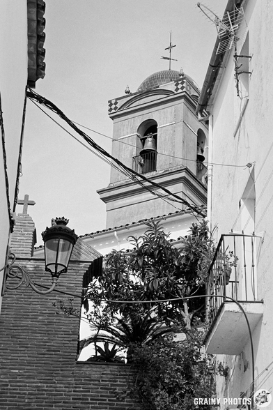 A charming black and white view of a narrow street featuring a bell tower, adorned with wires and a wall lantern, framed by lush greenery and architectural details of nearby buildings.