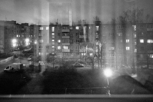 The black-and-white photo shows a close-up of a dark window with reflections of a room, which shows the courtyard and the house opposite with burning windows and lanterns.
