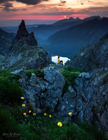 A woman is sitting on a mountain ridge above a lake, observing the sunset.