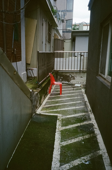 건물 사이 좁은 공간. 새빨간 플라스틱 의자 하나가 놓여있다. 하얀색 페인트로 빗금이 잔뜩 그어진 콘크리트 바닥에는 녹색 이끼가 잔뜩 끼어있다.